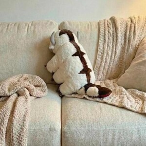 May include: A plush toy of a white and brown creature with horns, resembling a bison, sits on a beige couch. The toy has a brown stripe down its back and a red tail. A beige knitted blanket is draped over the couch.