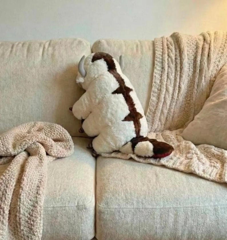 May include: A plush toy of a white and brown creature with horns, resembling a bison, sits on a beige couch. The toy has a brown stripe down its back and a red tail. A beige knitted blanket is draped over the couch.
