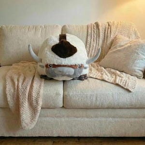 May include: A plush, round, white and brown stuffed animal with large, curved horns and a gray face. The toy is resting on a cream-colored couch with a matching blanket and pillow.