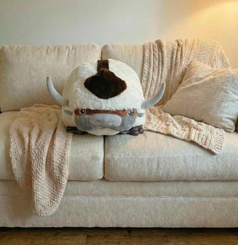 May include: A plush, round, white and brown stuffed animal with large, curved horns and a gray face. The toy is resting on a cream-colored couch with a matching blanket and pillow.