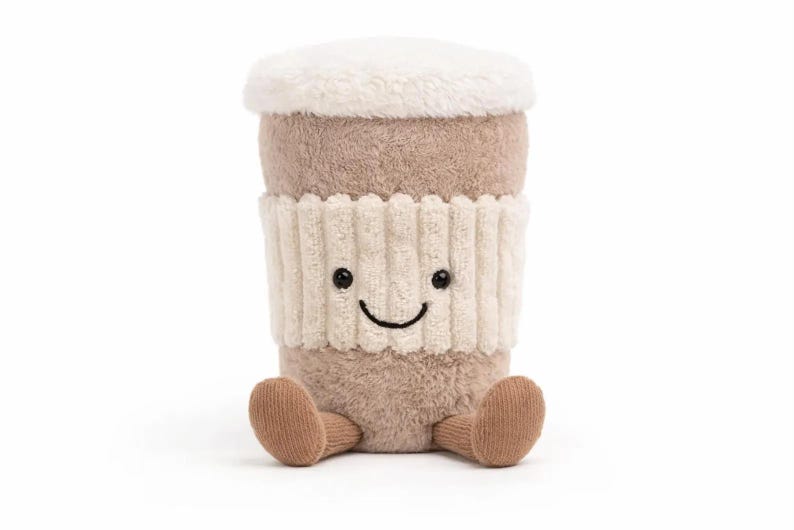 May include: A soft toy coffee cup with a smiling face, a brown body, and a cream-coloured lid. It has a cream-coloured ribbed sleeve and small brown feet. The toy is designed to sit upright.