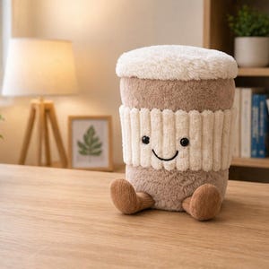 May include: A soft toy coffee cup with a smiling face, a brown body, a white ribbed sleeve, and a fluffy white top. The toy has small brown feet and is on a wooden surface. The background includes a lamp, plants, and a bookshelf.