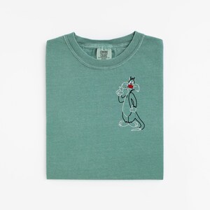 May include: A folded, seafoam green t-shirt with a small embroidered cartoon cat design on the front. The cat is outlined in black with red and white details. The shirt has a crew neck and a tag on the collar.