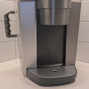 May include: A silver and black single-serve coffee maker. The machine has a water reservoir, a handle, and a drip tray. The top section is silver, and the front and base are black. The coffee maker is designed for making single cups of coffee.