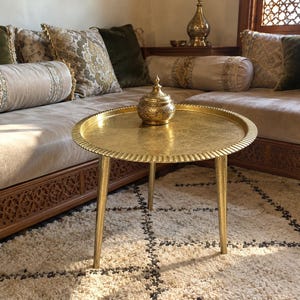 May include: A round gold-colored metal side table with three tapered legs and a textured, scalloped edge. A matching gold-colored metal lidded bowl with intricate engraved patterns sits in the center of the table surface.