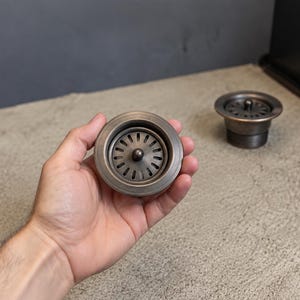 May include: A round, metallic sink drain strainer with a dark, brushed bronze finish. The strainer features a circular rim, a central knob, and a series of radiating slots for water drainage. A second identical strainer sits in the background.