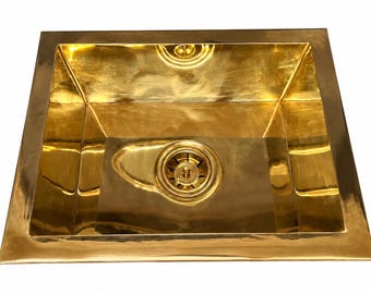 May include: A square kitchen sink made of polished brass with a reflective gold-toned surface. The basin features a wide, flat rim and a central circular drain assembly that matches the metallic finish of the sink body.