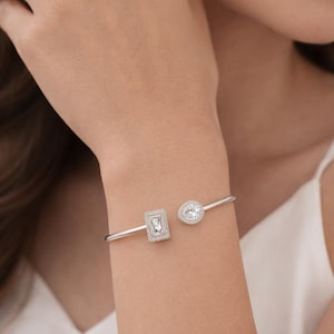 Puede incluir: Una pulsera de plata con dos elementos decorativos: una forma rectangular y un diseño en forma de pera, ambos incrustados con piedras claras y brillantes. La pulsera se lleva en la muñeca.