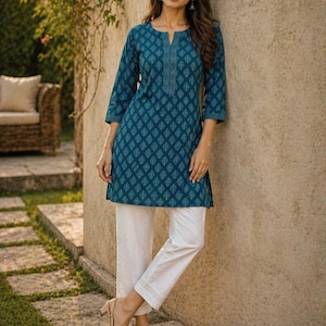 May include: Teal tunic with a geometric pattern and three-quarter sleeves paired with white cropped pants. The tunic has a split neckline and a detailed trim. The pants have a subtle hem detail. The outfit is completed with beige sandals.