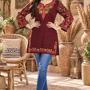 May include: A maroon tunic featuring intricate floral embroidery in shades of orange, pink, and green around the neckline, sleeves, and hem, paired with blue denim jeans and gold-toned pointed-toe flats. The tunic has long sleeves and a round neckline.