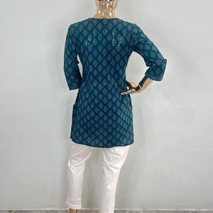 May include: A teal tunic with a leaf pattern and three-quarter sleeves, paired with white pants. The tunic has a round neckline and a slightly flared shape. The white pants have a lace trim at the ankles. The outfit is displayed on a mannequin.