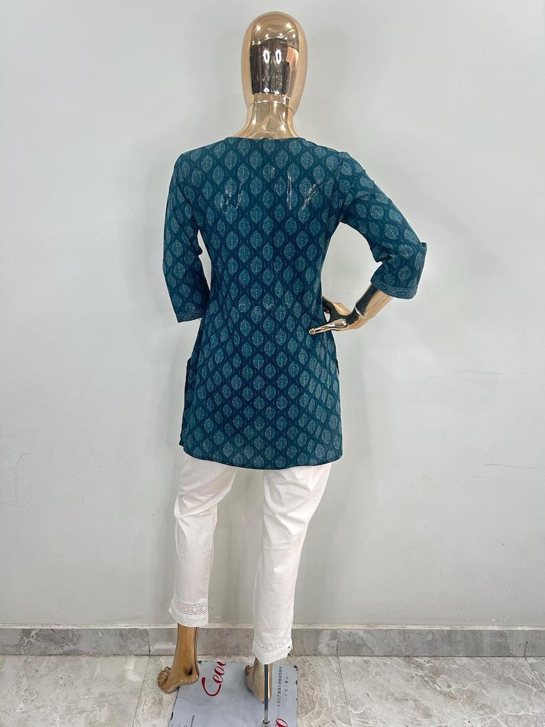 May include: A teal tunic with a leaf pattern and three-quarter sleeves, paired with white pants. The tunic has a round neckline and a slightly flared shape. The white pants have a lace trim at the ankles. The outfit is displayed on a mannequin.
