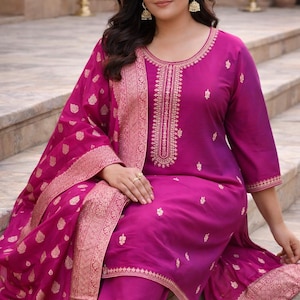 May include: A magenta-colored salwar kameez ensemble. The outfit features a tunic top with gold embroidery, matching pants, and a dupatta. The dupatta and the edges of the tunic are adorned with intricate gold patterns. The outfit is made of a shiny fabric.