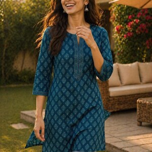 May include: A teal tunic with a leaf pattern and three-quarter sleeves. The tunic has a split neckline with embroidered detailing. The model is wearing white pants and silver earrings. The background includes greenery and outdoor furniture.