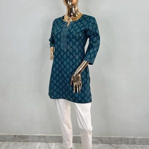 May include: Teal tunic with a leaf pattern and white pants. The tunic has a v-neck and three-quarter sleeves. The white pants have a lace trim at the ankles. The outfit is displayed on a gold mannequin.