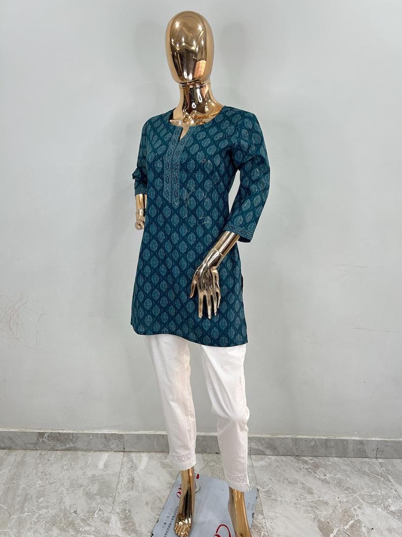 May include: Teal tunic with a leaf pattern and white pants. The tunic has a v-neck and three-quarter sleeves. The white pants have a lace trim at the ankles. The outfit is displayed on a gold mannequin.