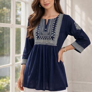 May include: A navy blue tunic top with intricate white embroidery around the neckline and sleeves. The top has a V-neck and three-quarter sleeves. The model is wearing white pants. The tunic top is a stylish addition to any wardrobe.