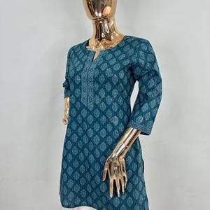 May include: Teal tunic with a leaf pattern and a V-neck. The three-quarter sleeves and the neckline are detailed with white embroidery. The tunic is paired with white pants. The mannequin has a gold-colored head and hands.