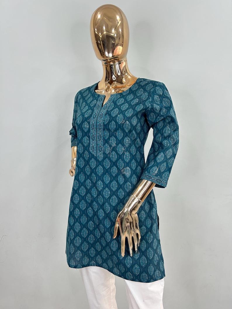 May include: Teal tunic with a leaf pattern and a V-neck. The three-quarter sleeves and the neckline are detailed with white embroidery. The tunic is paired with white pants. The mannequin has a gold-colored head and hands.