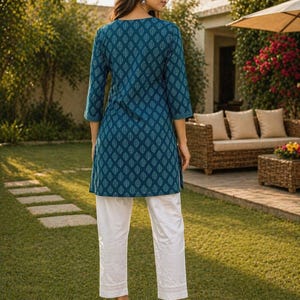 May include: A teal tunic with a leaf pattern and white pants. The tunic has three-quarter sleeves and a round neckline. The white pants have a straight leg and a lace trim at the hem. The outfit is worn with beige sandals.