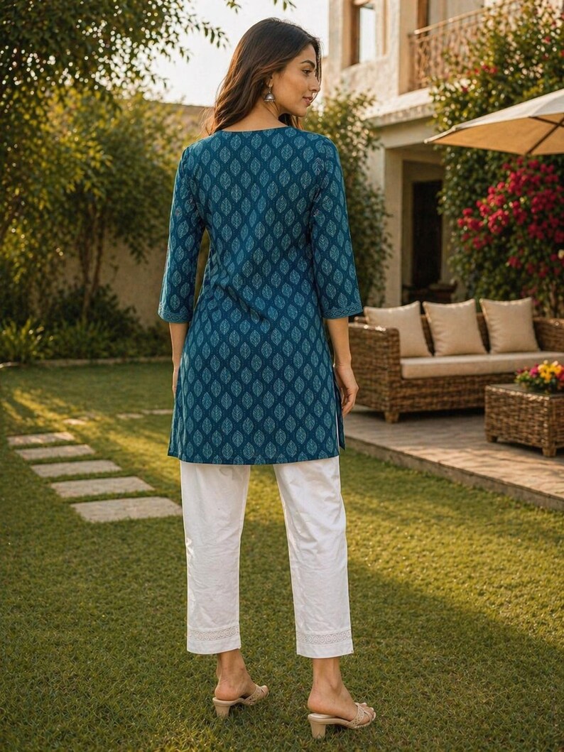 May include: A teal tunic with a leaf pattern and white pants. The tunic has three-quarter sleeves and a round neckline. The white pants have a straight leg and a lace trim at the hem. The outfit is worn with beige sandals.