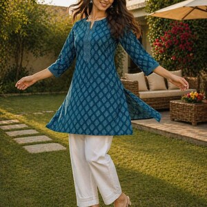 May include: A teal tunic with a leaf pattern and white pants. The tunic has three-quarter sleeves and a decorative neckline. The white pants are cropped and have a subtle embroidered hem. The outfit is worn with gold sandals.
