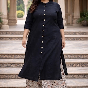 May include: A black tunic dress with a button-down front and three-quarter sleeves. The dress is worn with a patterned skirt. The model is wearing gold sandals. The background features a stone staircase and architectural details.