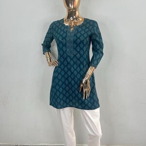 May include: A teal tunic with a leaf pattern and white pants. The tunic has a V-neck and three-quarter sleeves. The pants are straight-legged and have a decorative hem. The outfit is displayed on a mannequin.