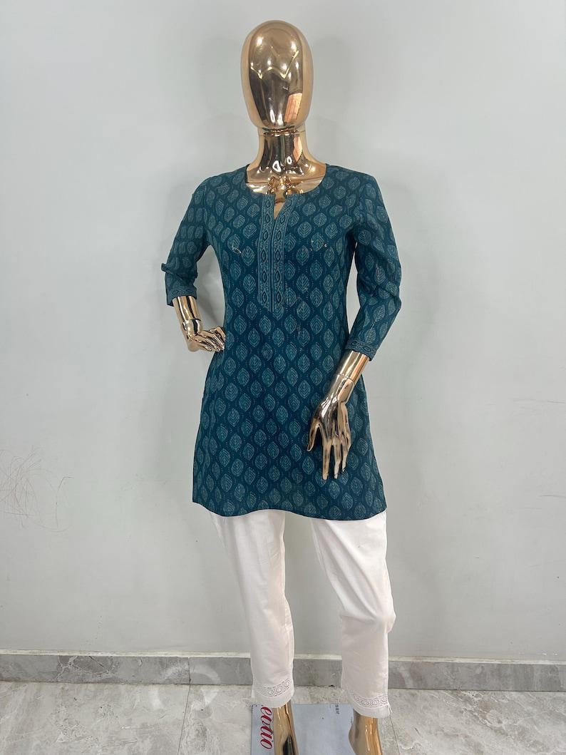 May include: A teal tunic with a leaf pattern and white pants. The tunic has a V-neck and three-quarter sleeves. The pants are straight-legged and have a decorative hem. The outfit is displayed on a mannequin.