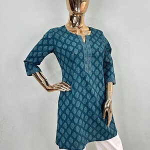 May include: Teal tunic with a leaf pattern and three-quarter sleeves. The tunic has a V-neck with white embroidered detailing and is paired with white pants. The mannequin has a gold-colored head and arms.