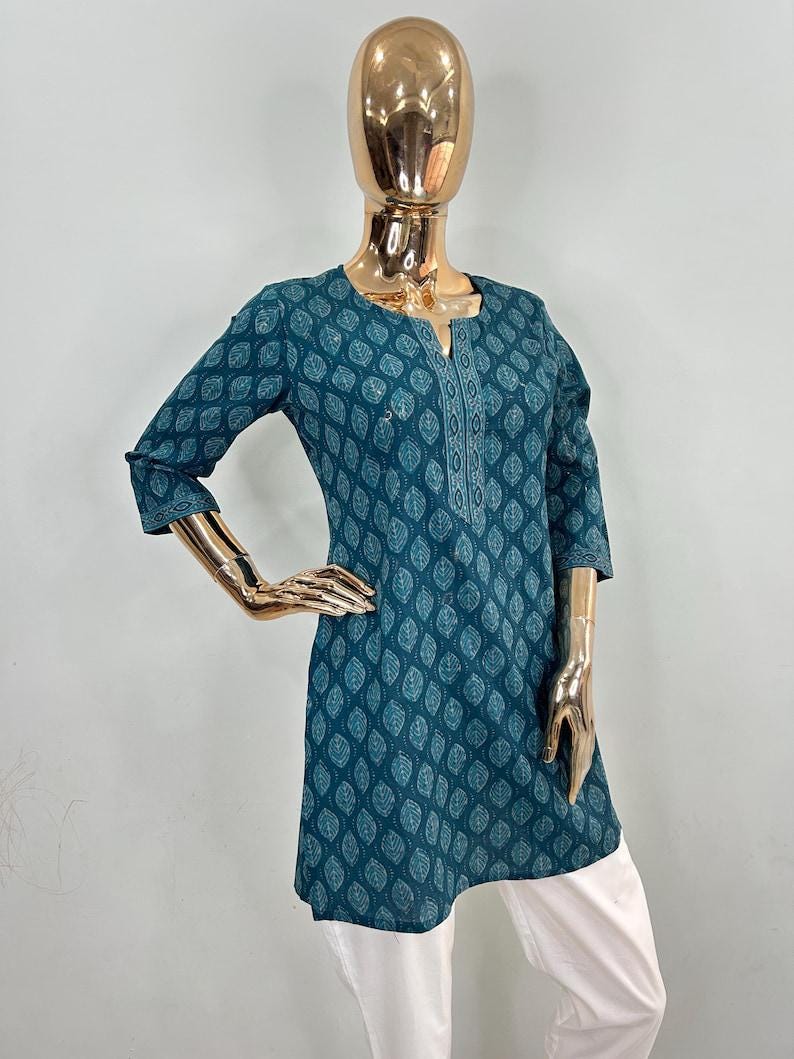 May include: Teal tunic with a leaf pattern and three-quarter sleeves. The tunic has a V-neck with white embroidered detailing and is paired with white pants. The mannequin has a gold-colored head and arms.