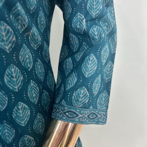 May include: Teal-colored fabric with a leaf pattern. The fabric is printed with white and light teal leaf shapes. The sleeve cuff has a decorative border. The garment is displayed on a gold mannequin arm.