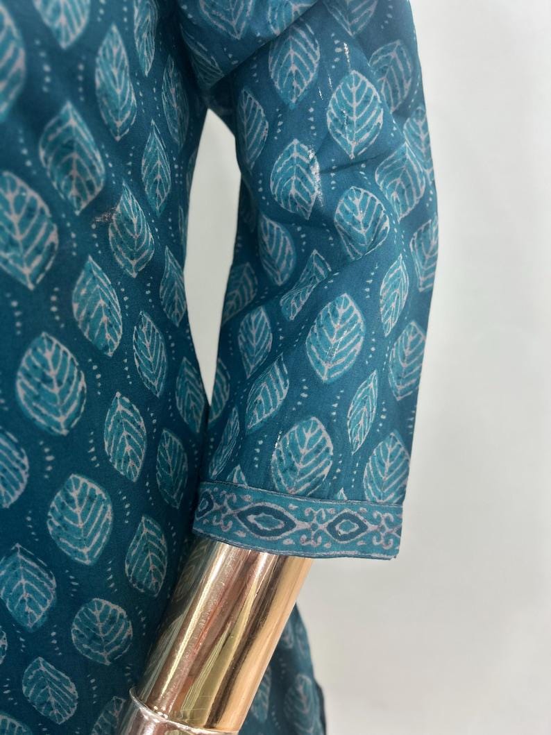 May include: Teal-colored fabric with a leaf pattern. The fabric is printed with white and light teal leaf shapes. The sleeve cuff has a decorative border. The garment is displayed on a gold mannequin arm.