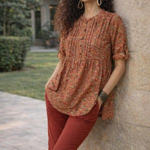 May include: A woman wearing a rust-colored outfit. The top is a short-sleeved blouse with a floral pattern in red, green, and tan. The pants are a matching rust color. The woman is leaning against a stone wall.