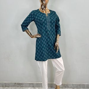 May include: A teal tunic with a leaf pattern and three-quarter sleeves paired with white pants. The tunic has a detailed neckline and a slightly flared shape. The white pants have a lace trim at the hem. The outfit is displayed on a gold mannequin.