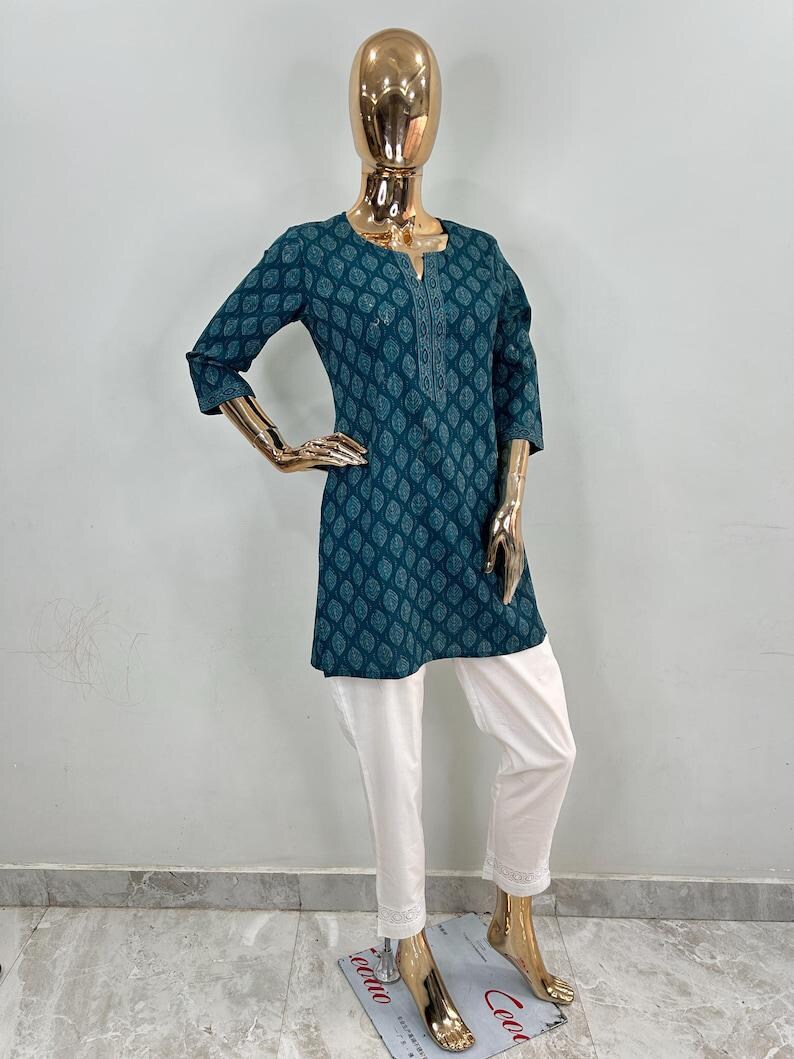 May include: A teal tunic with a leaf pattern and three-quarter sleeves paired with white pants. The tunic has a detailed neckline and a slightly flared shape. The white pants have a lace trim at the hem. The outfit is displayed on a gold mannequin.