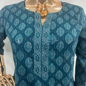 May include: Teal-colored tunic with a leaf pattern. The tunic features a V-neckline with a decorative border and three-quarter sleeves. The leaf design is in a lighter shade of teal, creating a contrast against the darker background.