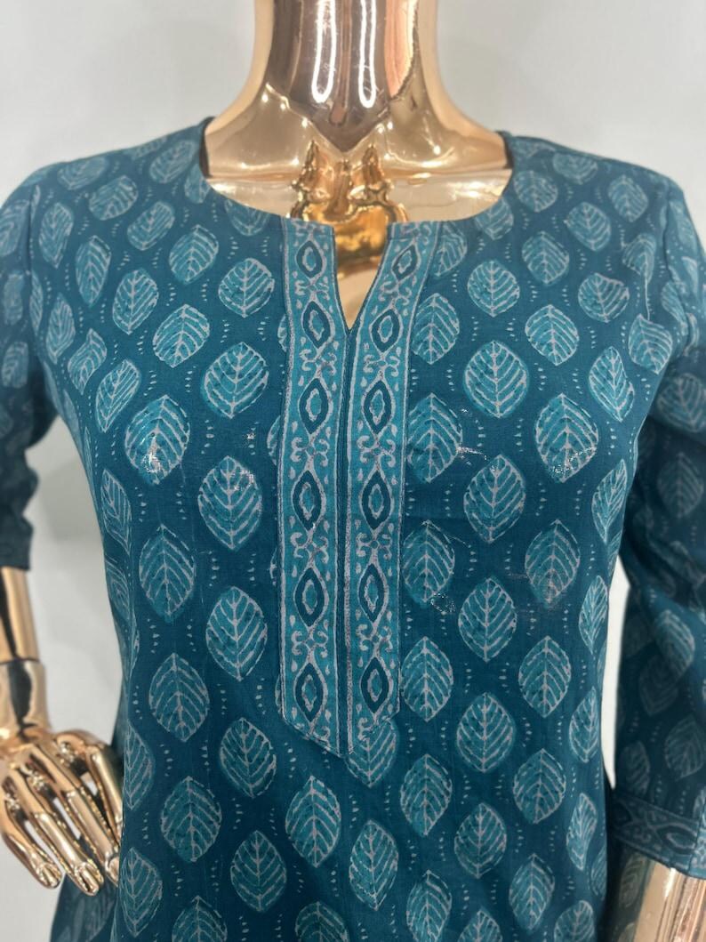 May include: Teal-colored tunic with a leaf pattern. The tunic features a V-neckline with a decorative border and three-quarter sleeves. The leaf design is in a lighter shade of teal, creating a contrast against the darker background.