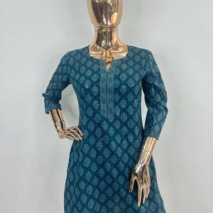 May include: Teal tunic with a leaf pattern, displayed on a mannequin. The tunic has a square neckline, three-quarter sleeves, and a decorative embroidered panel down the front. The tunic is paired with white pants.