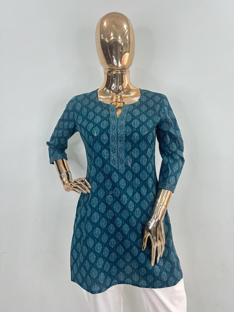 May include: Teal tunic with a leaf pattern, displayed on a mannequin. The tunic has a square neckline, three-quarter sleeves, and a decorative embroidered panel down the front. The tunic is paired with white pants.