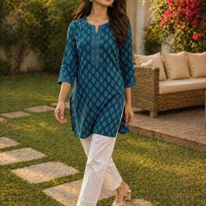 May include: A woman wearing a teal tunic with a white leaf pattern and white pants. The tunic has a split neckline and three-quarter sleeves. The pants are ankle-length. She is wearing sandals and walking on stone pavers in a garden.
