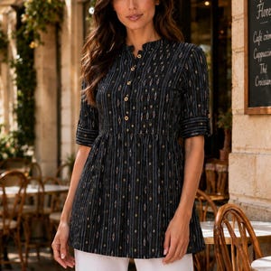 May include: A black tunic top with a vertical striped pattern in gold, white, and grey. The top has a mandarin collar, button-down front, and three-quarter sleeves. The model is wearing white trousers. The background shows a cafe setting.