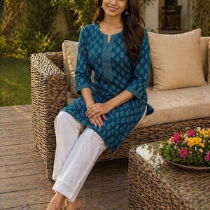 May include: A woman wearing a teal kurta with a leaf pattern and white pants. The kurta has a split neckline and three-quarter sleeves. The pants have a lace trim at the hem. She is sitting on a wicker sofa.