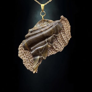 Op de afbeelding: Een hangerketting met een goudkleurige ketting en een groot, onregelmatig middelpunt. Het centrale deel combineert gladde, donkerbruine, gemarmerde hars met gouden glitteraccenten en een gestructureerde, koperkleurige gehaakte stoffen rand.