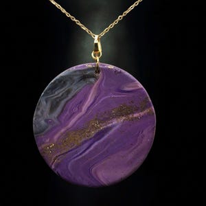 Op de afbeelding: Een ketting met een ronde hanger en een gouden ketting. De hanger heeft een wervelend ontwerp van paars, zwart en gouden glitter. De ketting staat tegen een zwarte achtergrond.