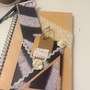 Puede incluir: Un diario hecho a mano con una cubierta con estampado de cebra, adornado con una etiqueta decorativa que dice "Notes & Dreams". El diario está adornado con encaje, perlas de imitación y flores de papel.