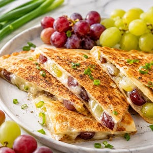 Puede incluir: Un plato de quesadillas rellenas de queso fundido y uvas, adornadas con cebolletas. Las quesadillas están cortadas en triángulos y dispuestas en un plato blanco, junto a racimos de uvas rojas y verdes.