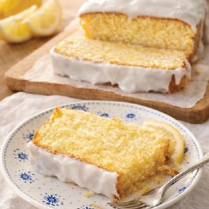Puede incluir: Una rebanada de pastel de limón con glaseado blanco sobre un plato con estampado azul y blanco, con un tenedor. Un pan de pastel de limón con una rebanada cortada, sobre una tabla de cortar de madera, con rodajas de limón.