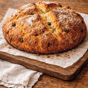 Puede incluir: Un pan redondo recién horneado, con corteza dorada y pasas oscuras, descansa sobre papel de hornear en una tabla de madera. El pan está ligeramente espolvoreado con harina, lo que le da un aspecto rústico y casero.