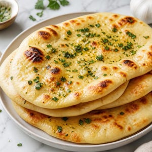 May include: A stack of three golden-brown naan flatbreads on a white plate, topped with melted butter, minced garlic, and fresh green cilantro. The bread features charred spots from cooking and is served with whole garlic cloves nearby.